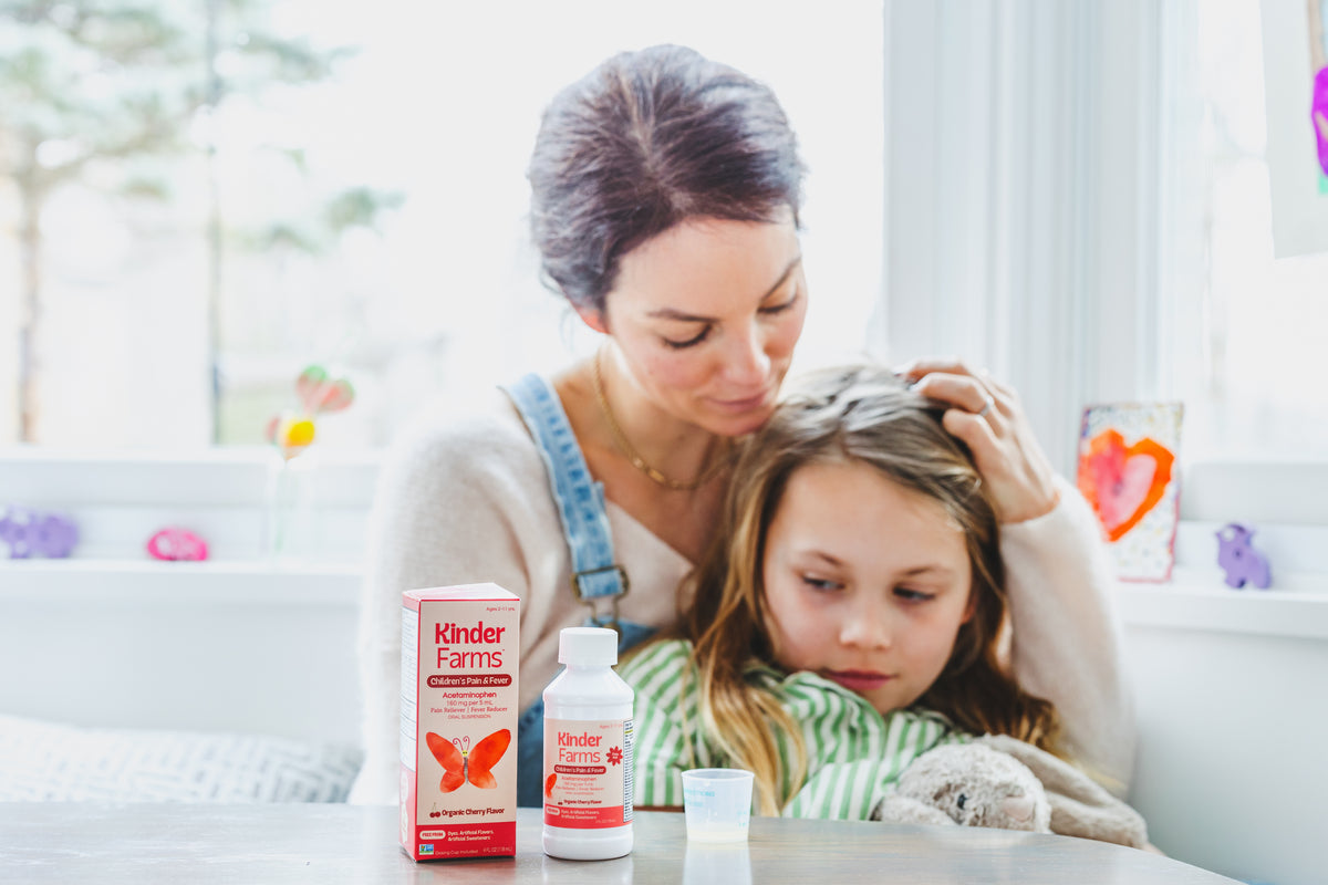 Mother comforting sick daughter. A bottle of KinderFarms Pain and Fever is on a counter