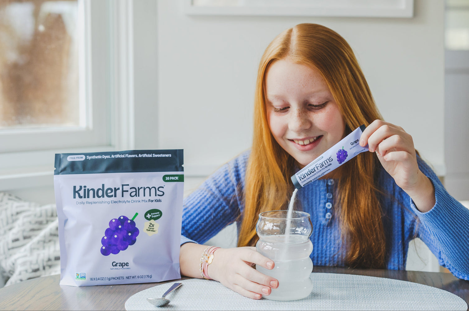 A girl with red hair smiles while pouring a KinderFarms Grape Electrolyte Drink Mix packet into a glass of water.