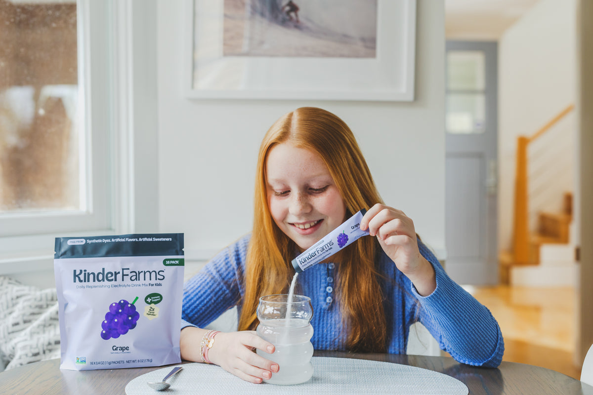A girl with red hair smiles while pouring a KinderFarms Grape Electrolyte Drink Mix packet into a glass of water.