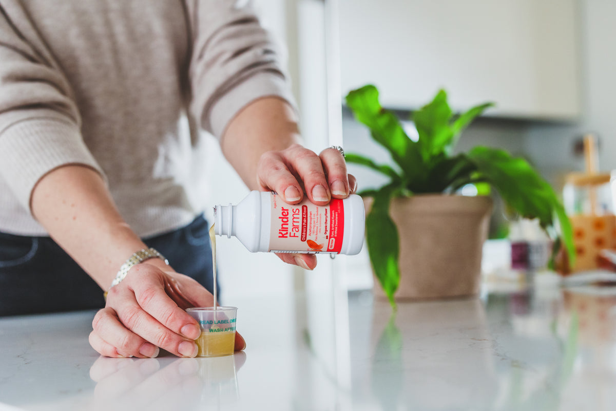 A person in a beige sweater pours KinderFarms from a white bottle with a red label into a small clear cup on a kitchen counter
