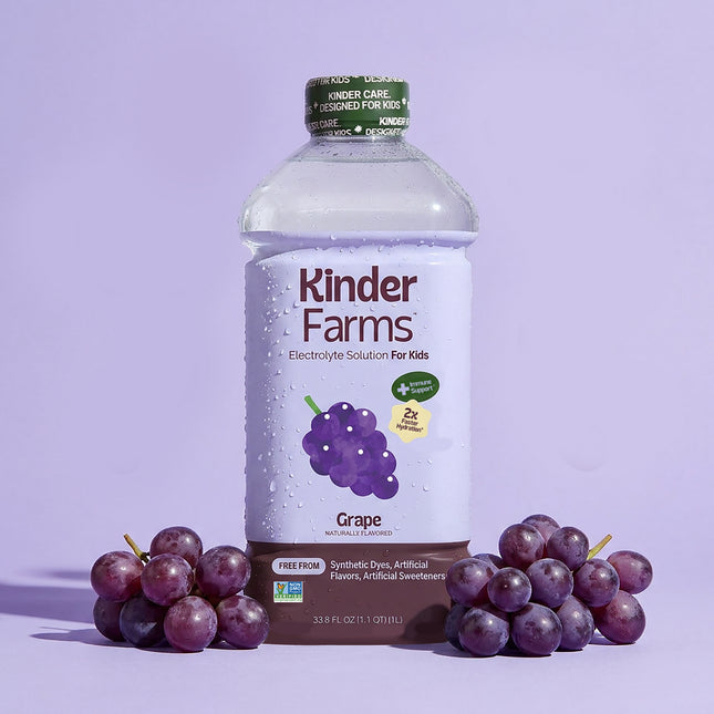 Kinder Farms Grape Electrolyte Solution bottle for kids standing between two clusters of fresh purple grapes against a light purple backdrop.