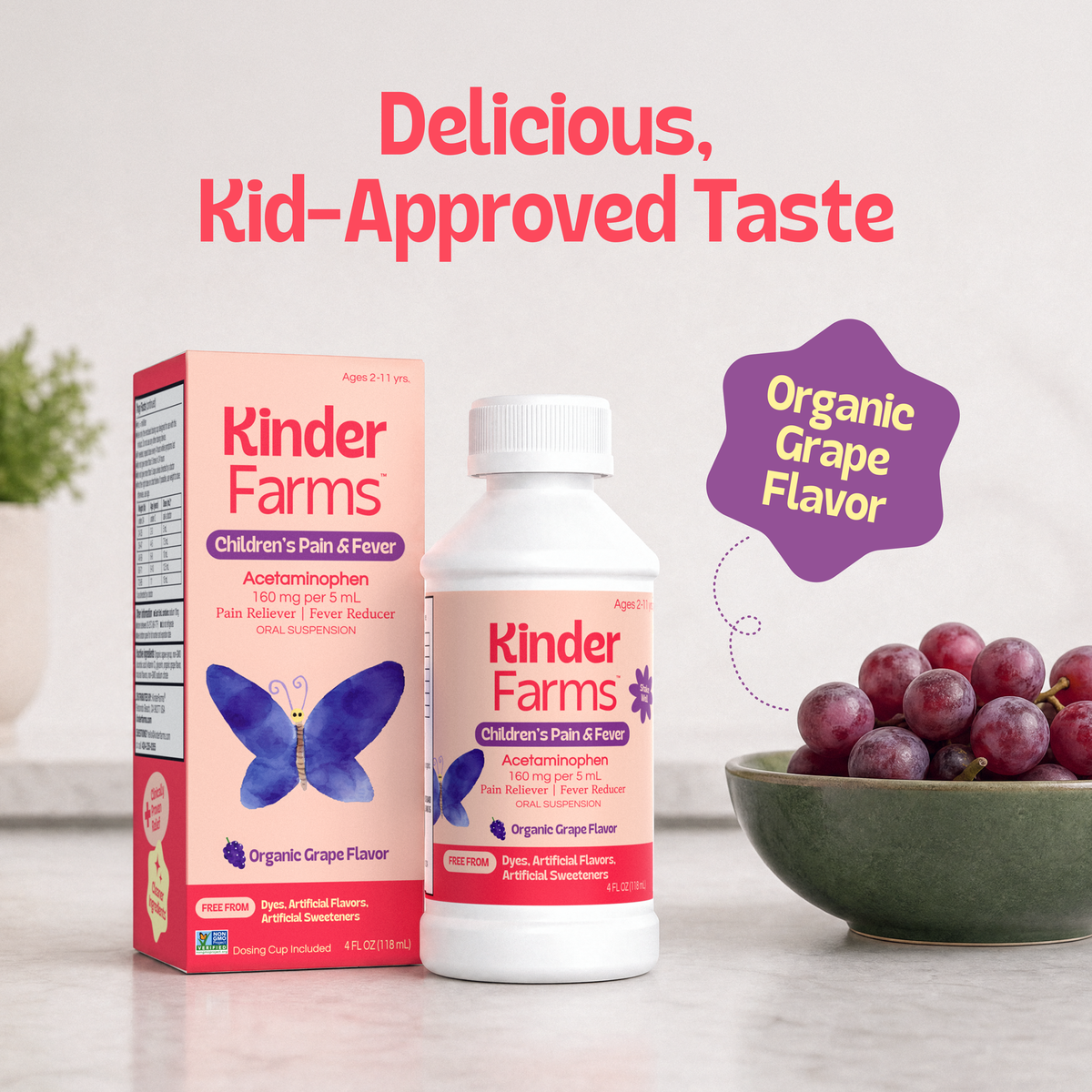 Kinder Farms Children's Pain & Fever liquid remedy shown with its retail box and bottle on a light surface, alongside a bowl of red grapes and the text 'Delicious, Kid-Approved Taste' and 'Organic Grape Flavor'. (Item 14 of 15)