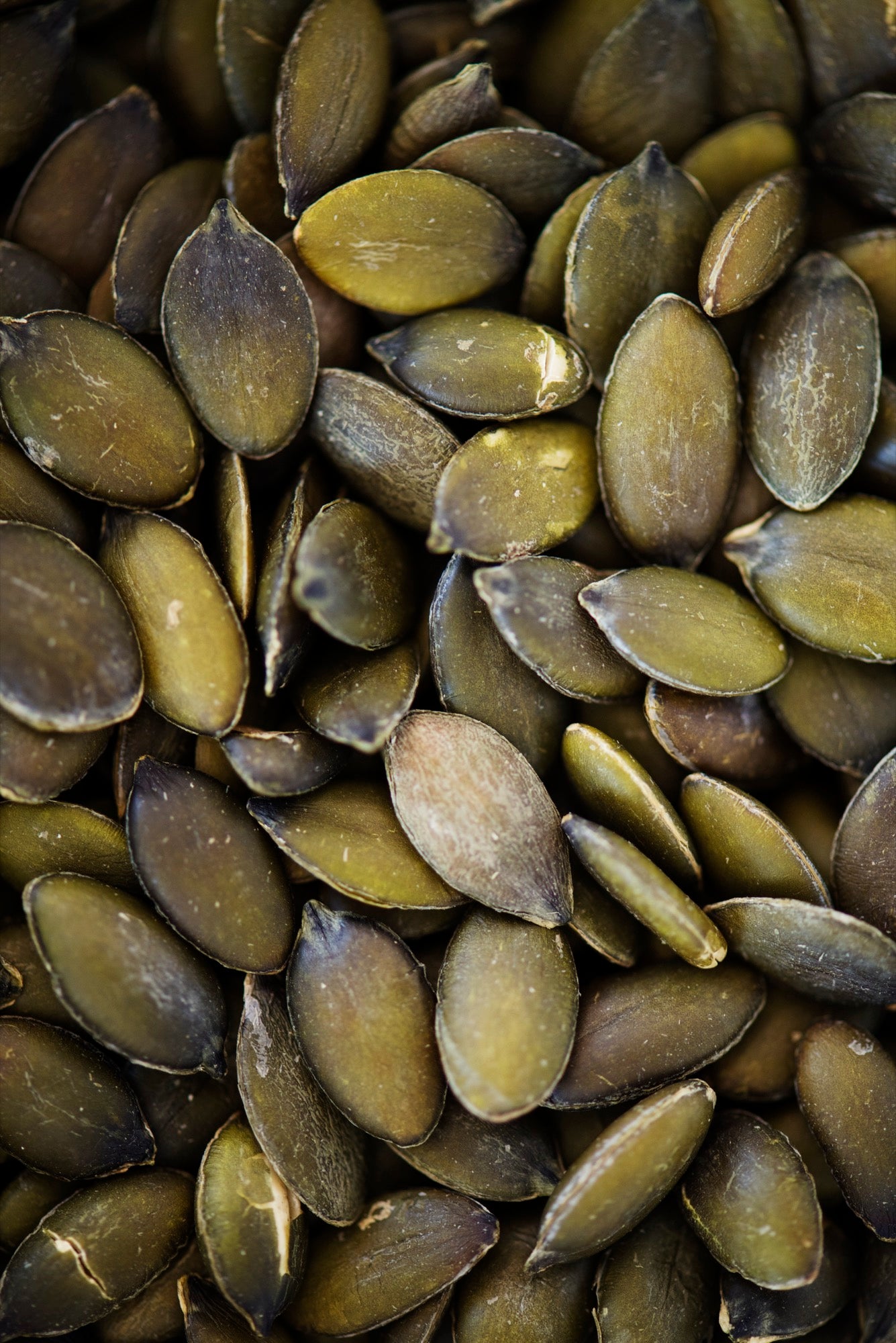 A close-up pile of raw pumpkin seeds with olive green and brown shells filling the entire frame.