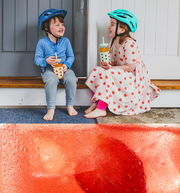 A boy in a blue helmet and a girl in a teal helmet sit on a doorstep with orange drinks, sharing a joyful moment.